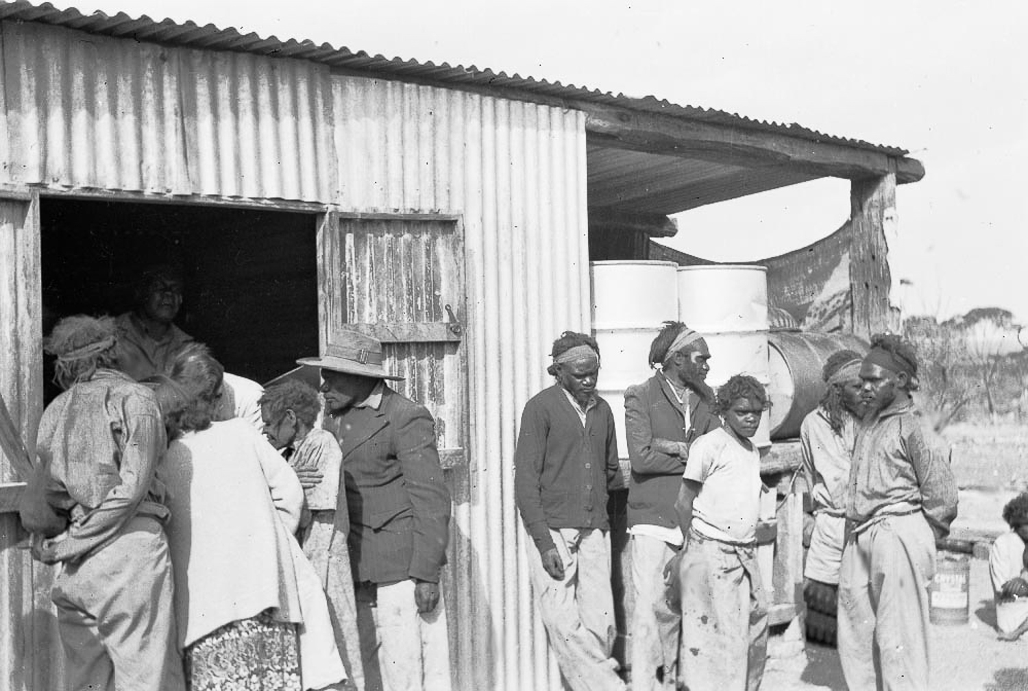 Jack Jamieson, Albert Jamieson, Frank Hogan, Arthur Jamieson and Bill Jamieson stand huddled together on the right, newcomers to the mission at Cundeelee. While the forced relocation of Anangu saw them dislocated from their homelands, descendants are grateful to the trackers who located them, as they were saved from potential disaster from the nuclear testing and a prolonged draught in Spinifex Country.
