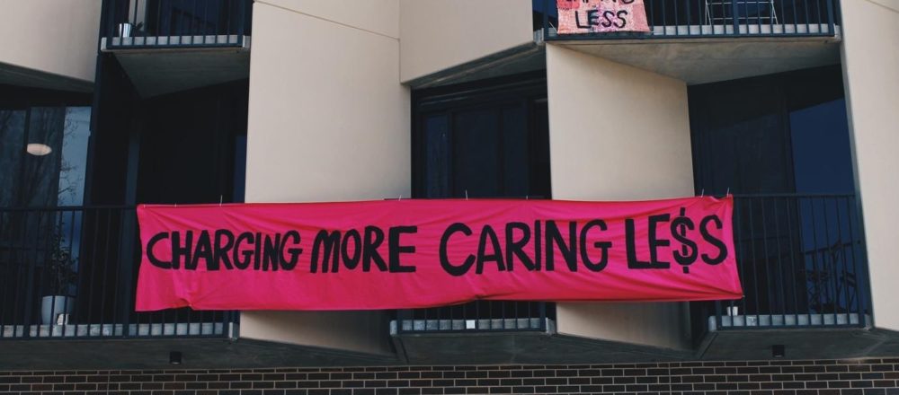 Black and cream building with grey bricks and a pink banner in the centre that reads "Charging more caring less"