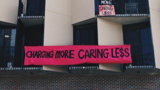Black and cream building with grey bricks and a pink banner in the centre that reads "Charging more caring less"